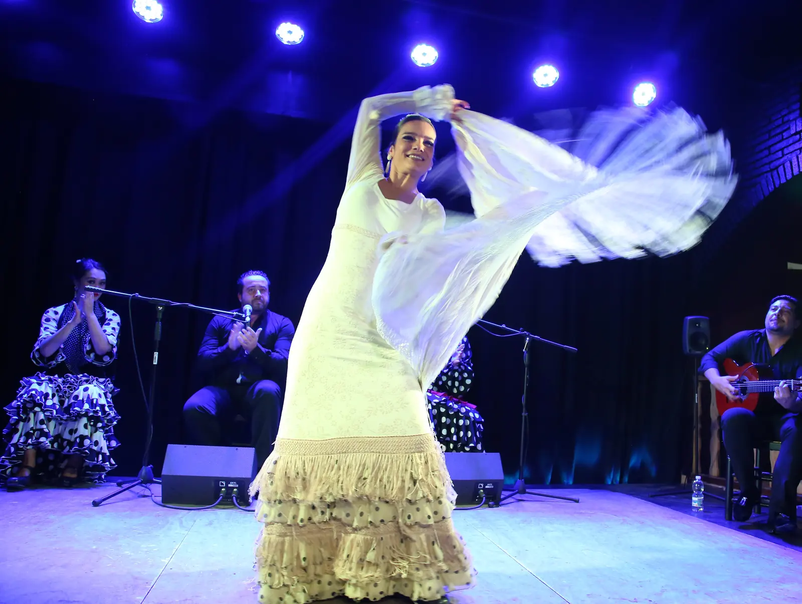 Flamenco tanečnice v bílých šatech a šátku s třásněmi uprostřed obratu na modře osvětleném pódiu, za ní sedí kytarista a umělci tleskající