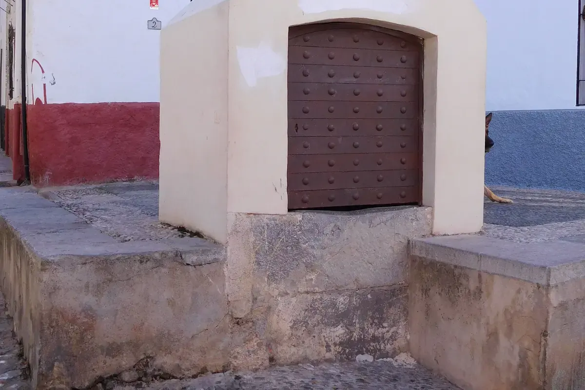 Eine weiß getünchte steinerne Zisterne mit einer beschlagenen Holztür in der Calle Panaderos im Albaicín, neben einer rot gestrichenen Mauer und einer steinernen Stufe